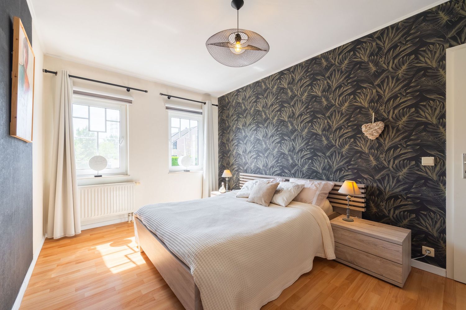 Cozy bedroom with dark floral accent wall, wooden floor, large bed with neutral bedding, two nightstands, and a bright window.