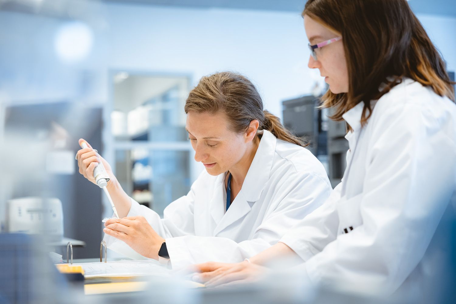 Two female scientists in white lab coats working with a pipette in a modern laboratory.