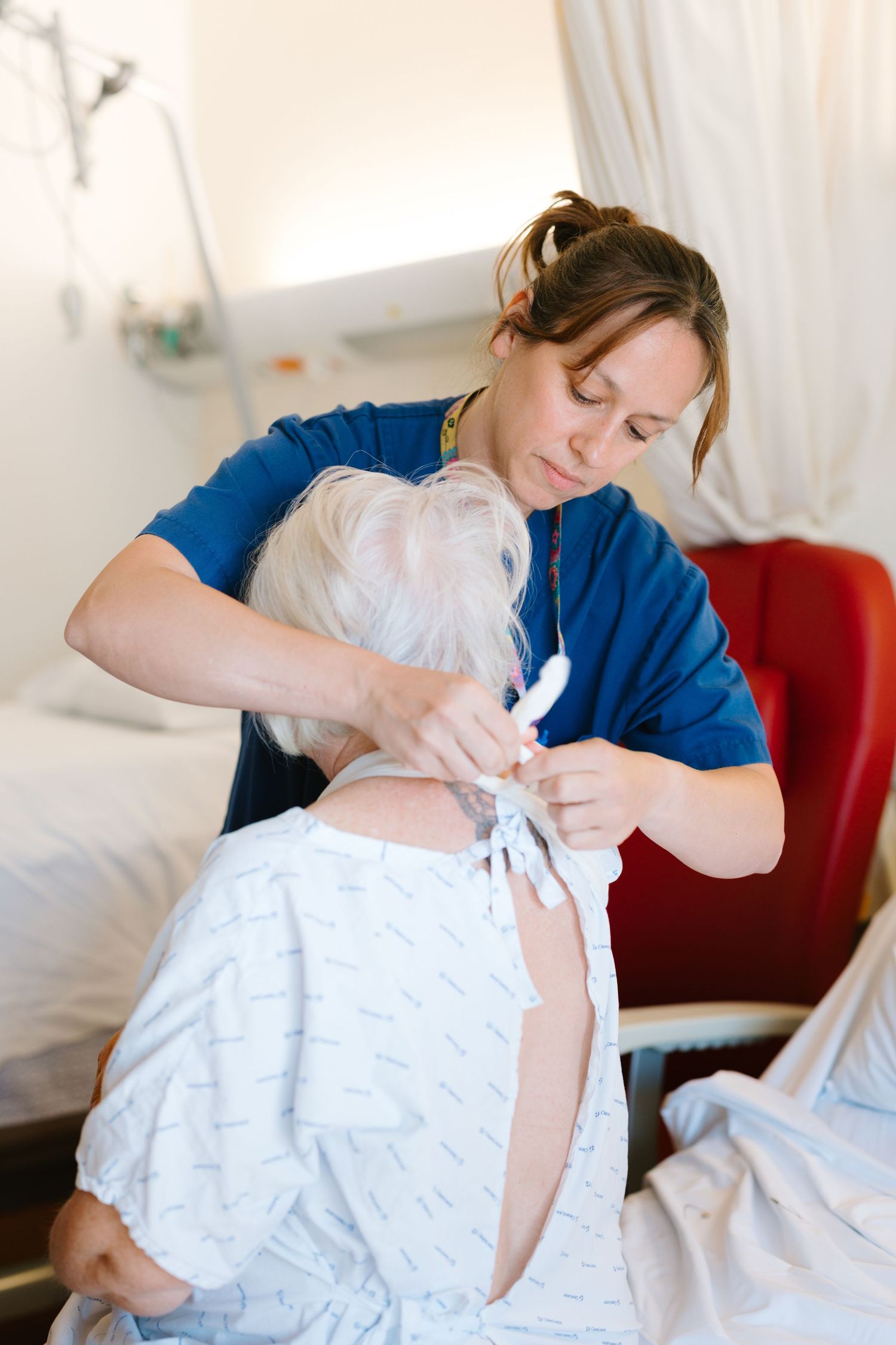 Nurse in blue scrubs tying hospital gown on elderly patient seated in medical facility.