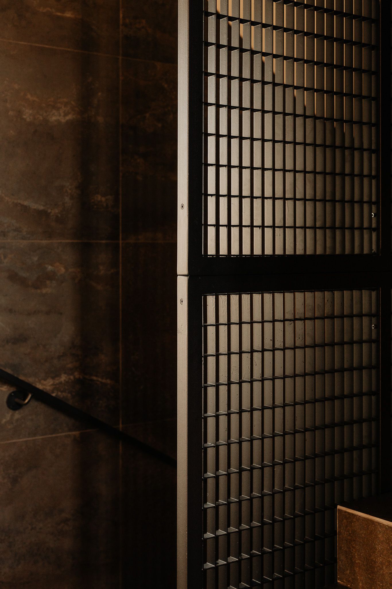 Dark moody stairwell featuring a metallic grid panel beside dark stone tiles and a black handrail.