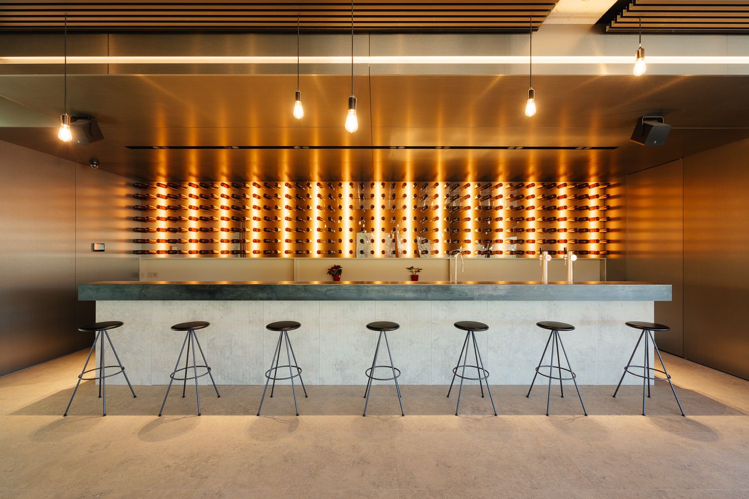Stylish bar with backlit wine bottle wall display, concrete counter, and industrial pendant lighting.