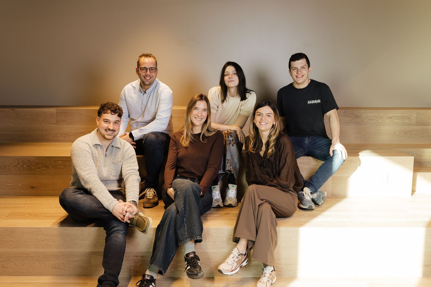 Group of seven smiling colleagues posing together on wooden steps in a modern office space.