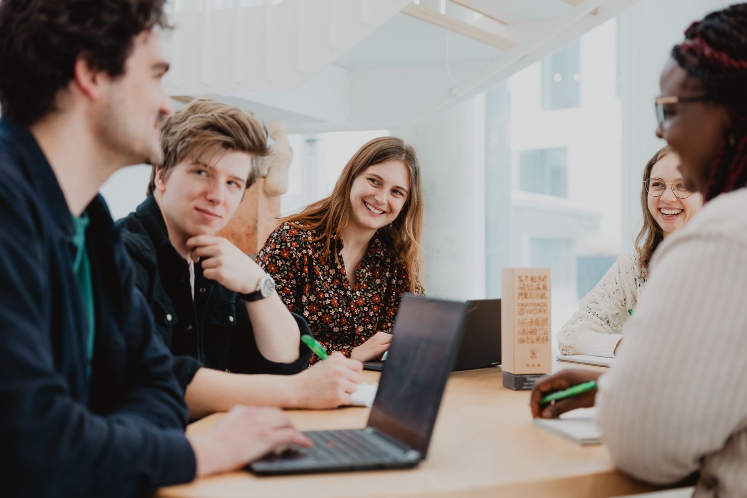 Diverse group of young professionals laughing and collaborating around a table with a laptop in a bright office.