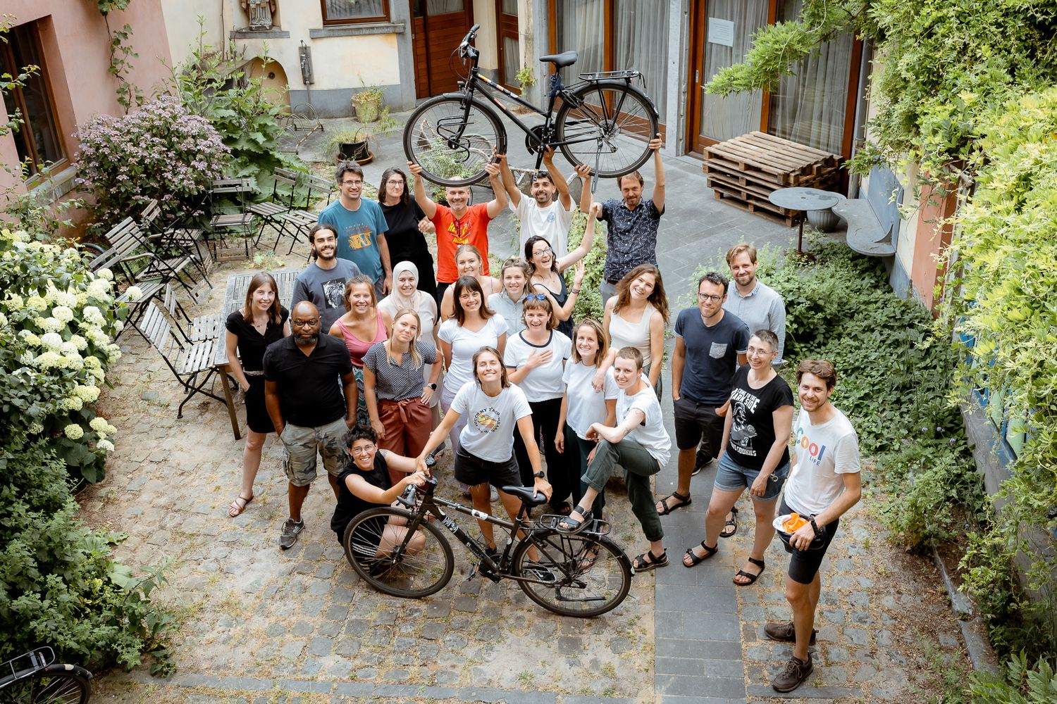 Large group of cyclists and team members posing together in a courtyard, holding bicycles overhead in celebration.