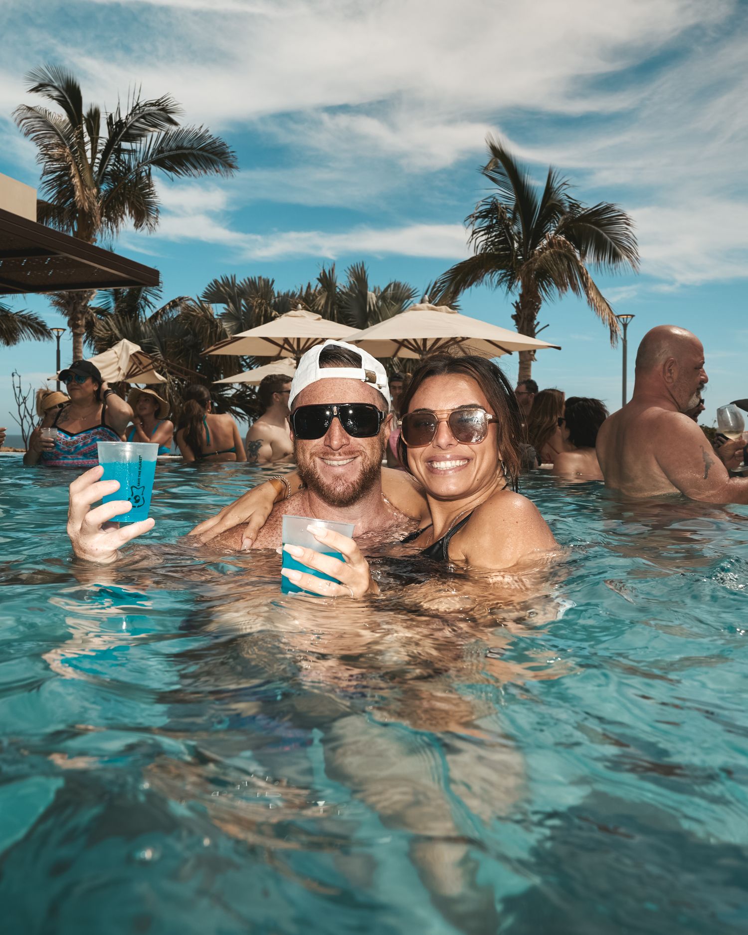 Smiling couple in the pool holding drinks at a daytime pool party, relaxed candid event photo.