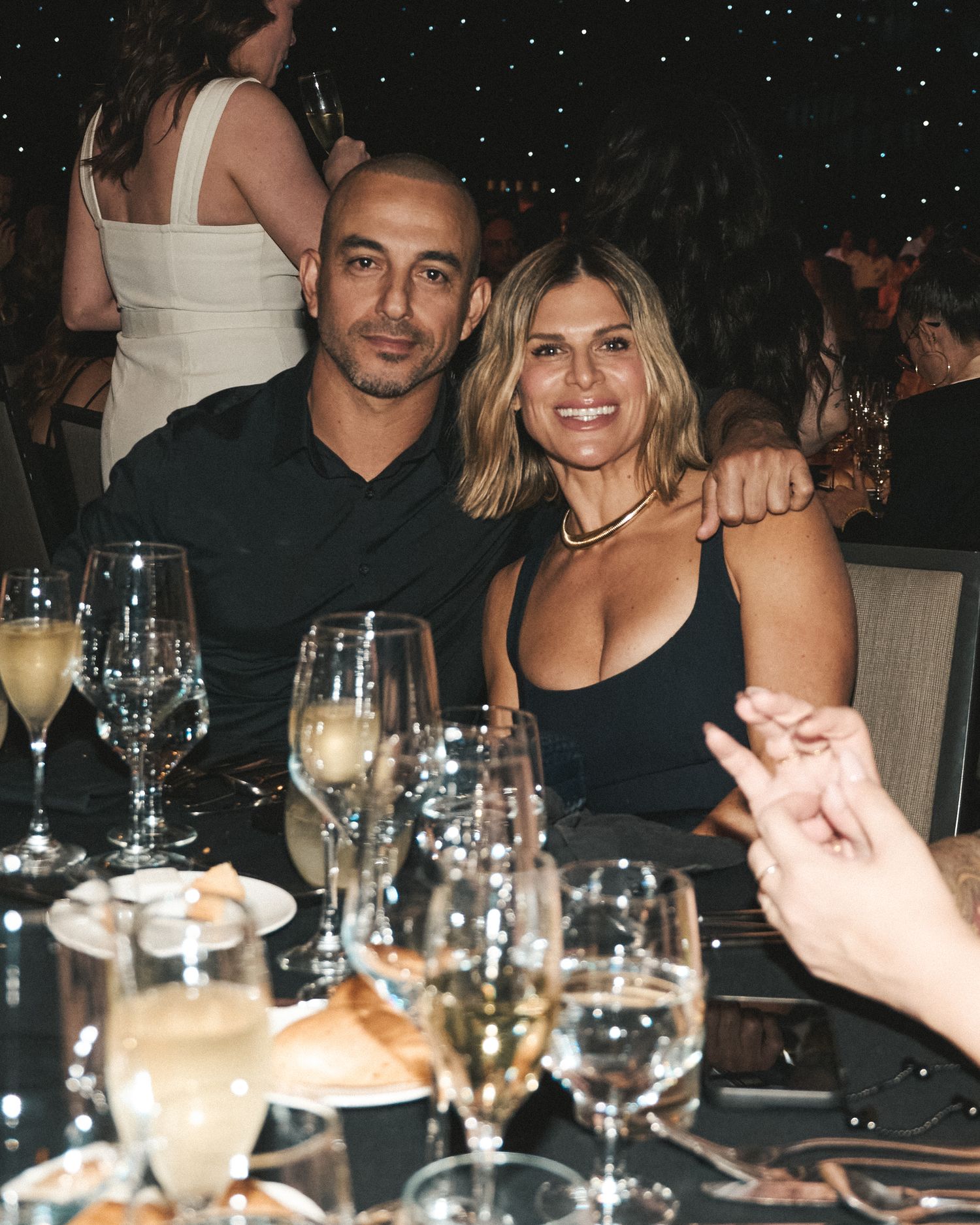 Couple seated at a decorated gala dinner table, smiling with wine glasses and place settings in front of them.
