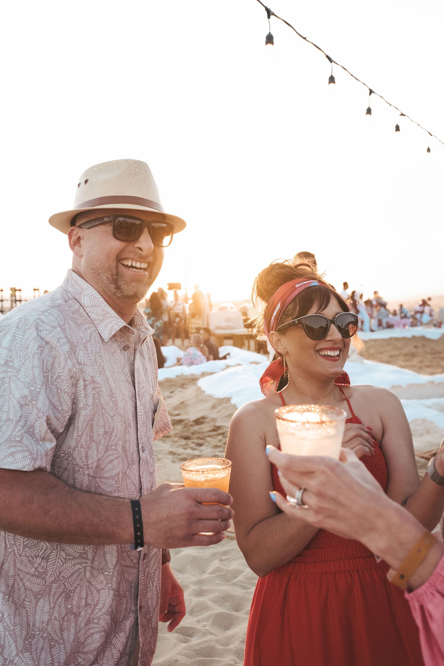 Couple laughing with drinks at a beach party at sunset, stylish editorial event photography.