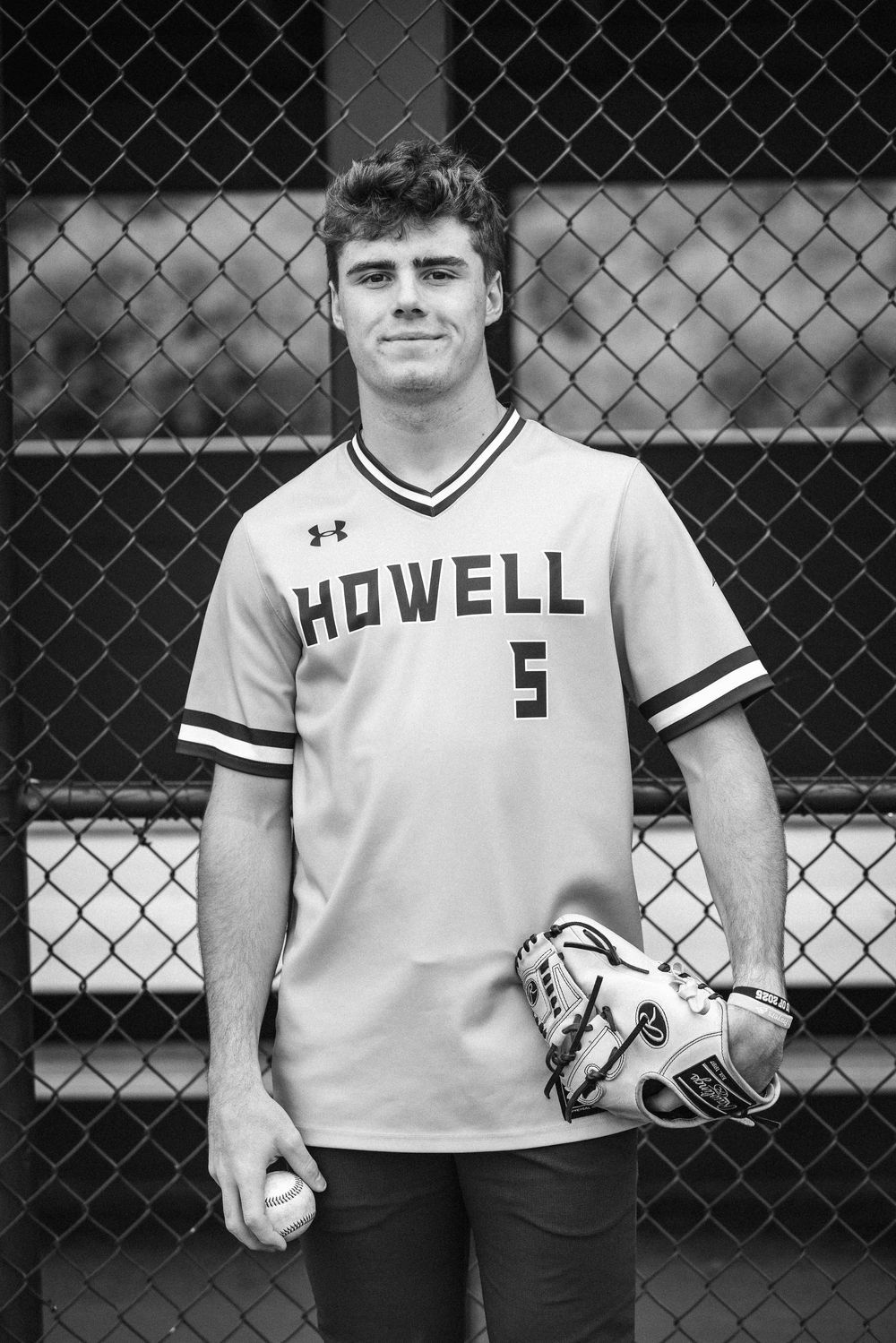 Editorial black and white senior portrait with strong tonal contrast.” Senior pic in Howell baseball jersey standing by a chain-link fence.