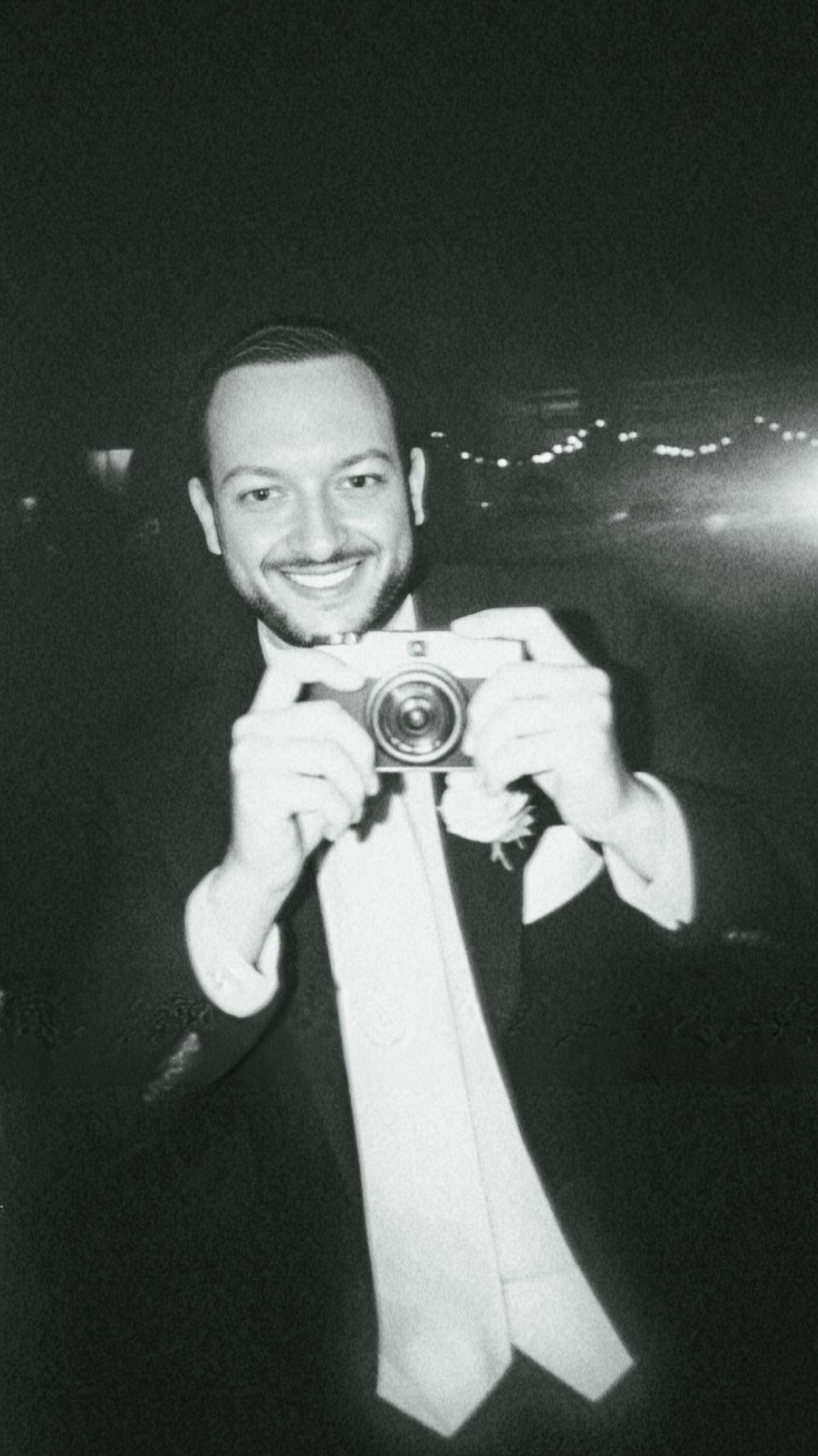 A vintage-style black and white photo of a person in formal wear holding a camera and smiling. Lifestyle photographer in O'Fallon, Missouri.