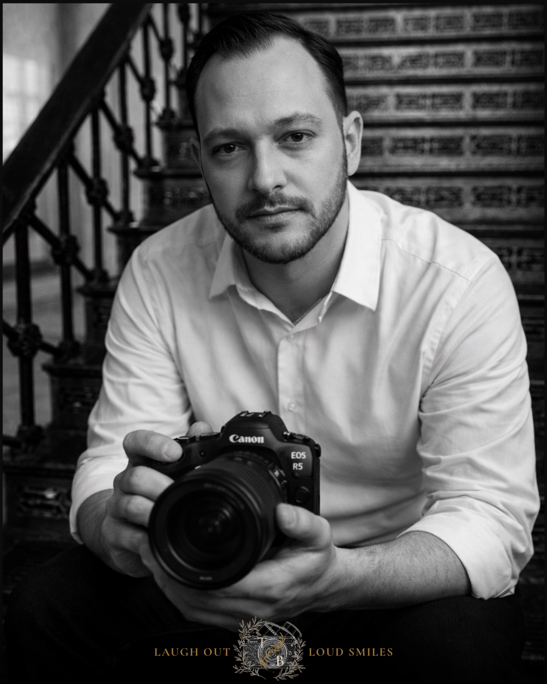 Black and white portrait of a male photographer holding a Canon EOS camera, seated near ornate staircase railings.
