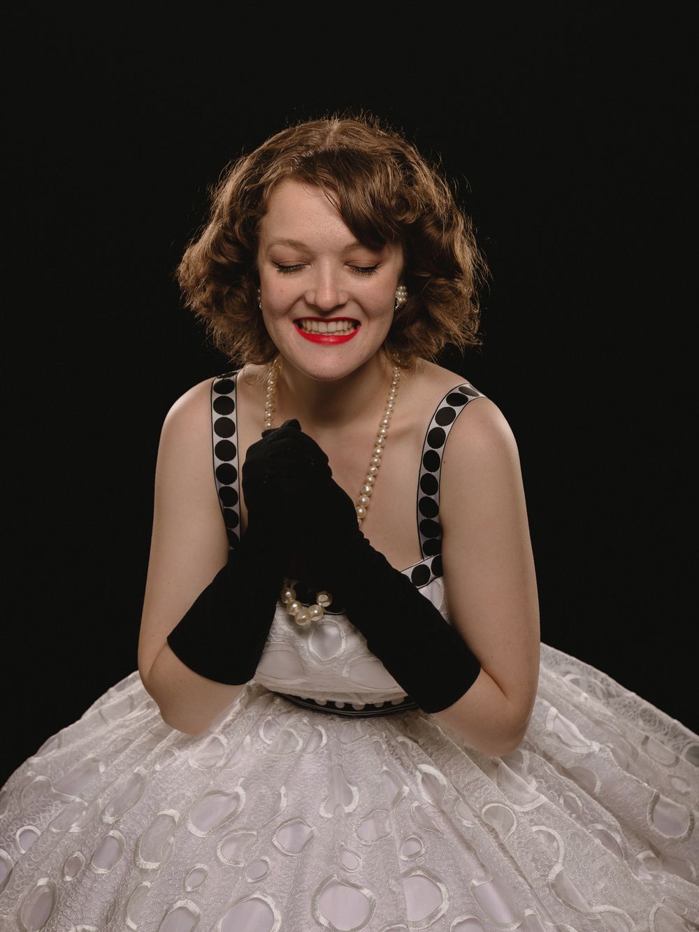 Model session in Saint Ann, Mo of a person in black gloves and a white dress with circular patterns poses against a dark background. Pin-up