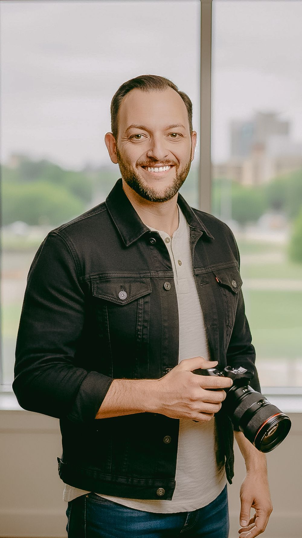 Laugh Out Loud Smiles, Saint Charles County, Missouri Photographer in black denim jacket holding camera in bright indoor setting. Headshot