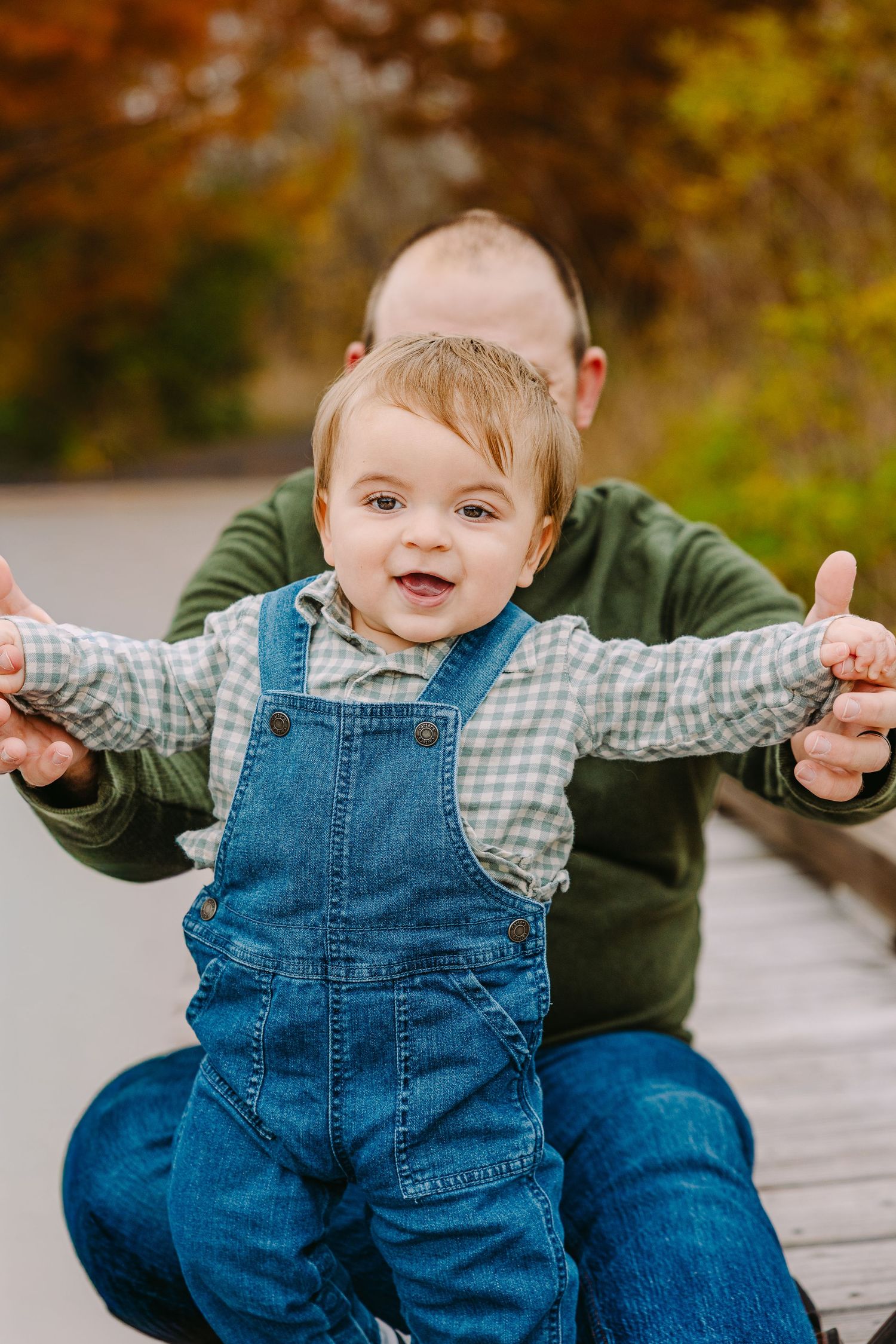 Kid Portrait in O'Fallon, MO ofchild wearing denim overalls and a checkered shirt smiles while being supported on an outdoor deck in autumn.
