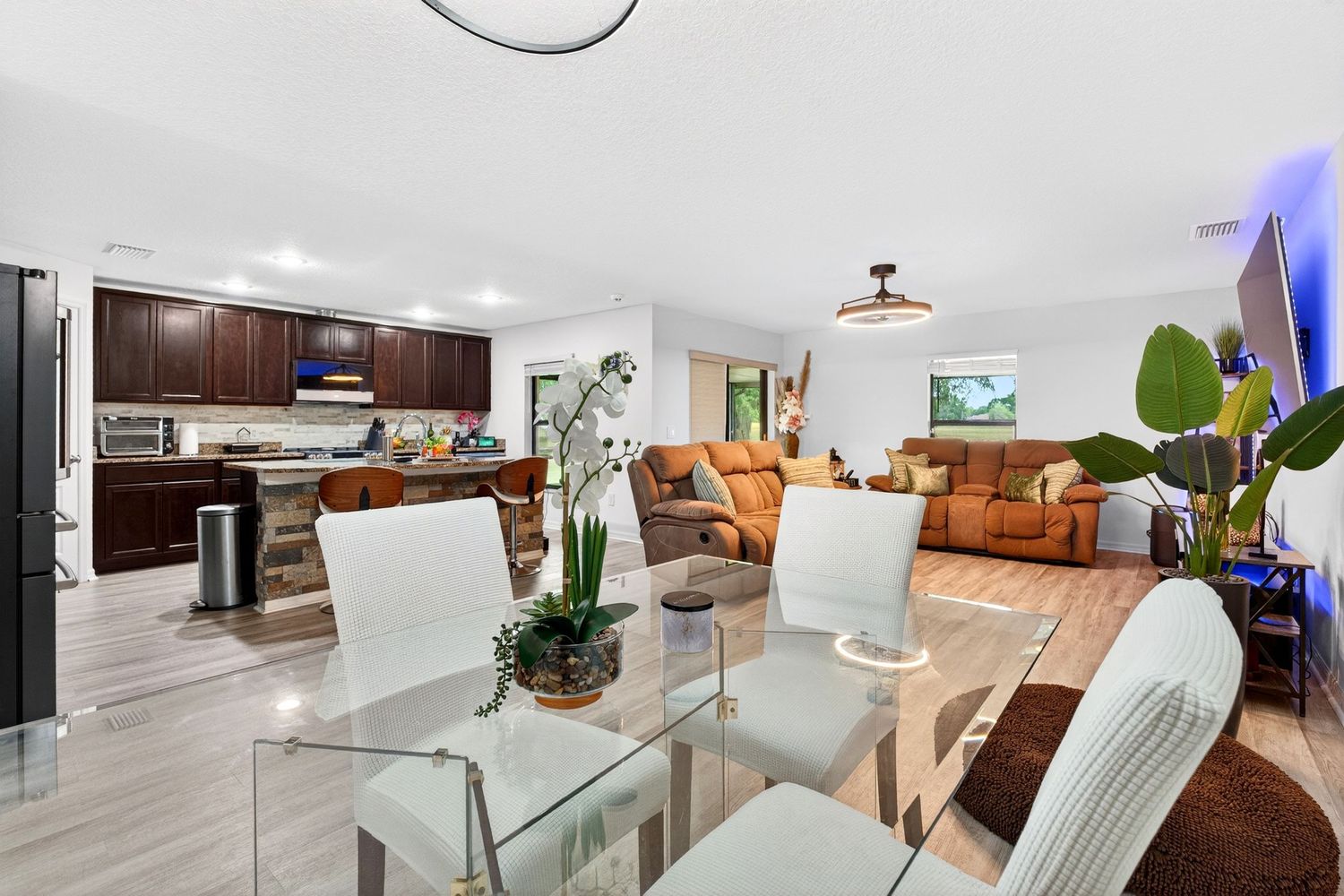 Open-concept dining area with glass table, white chairs, dark wood kitchen cabinets, and cozy living room in background.