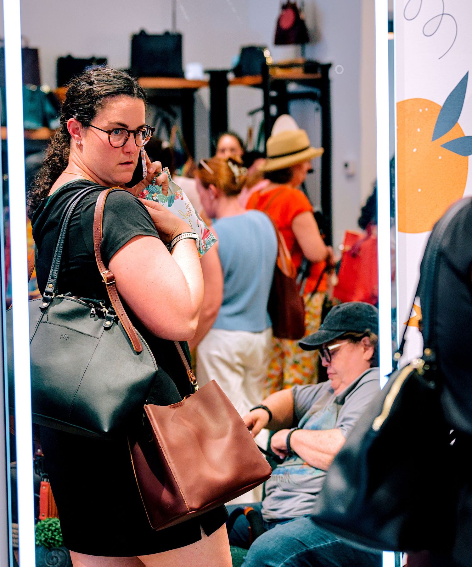 Woman with glasses and brown handbag browsing a busy retail shop with other shoppers in the background.