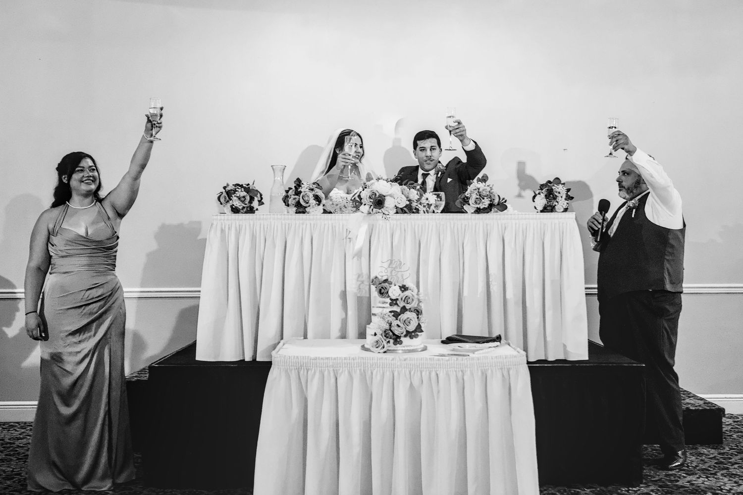 Black and white wedding reception toast with bride, groom, and guests raising glasses at decorated head table.