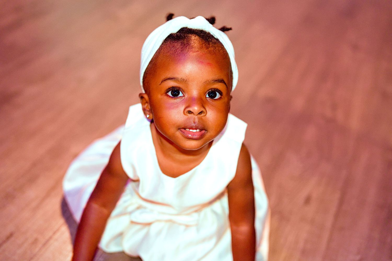 Adorable Black baby girl in white dress and headband looks up while crawling on hardwood floor.