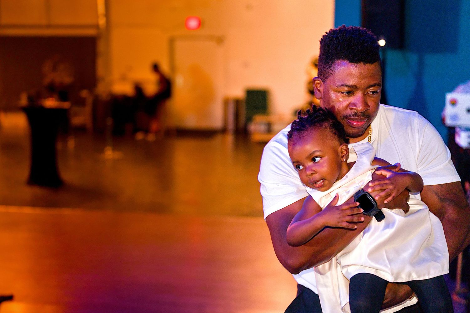 A man in a white shirt crouches down hugging a young child on a warmly lit event dance floor.