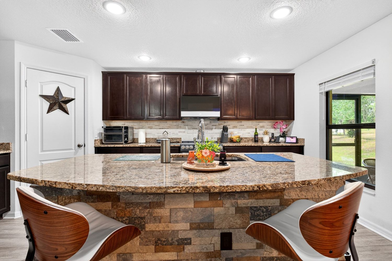 Modern kitchen with dark wood cabinets, granite island, bar stools, stainless appliances, and natural light from large window.