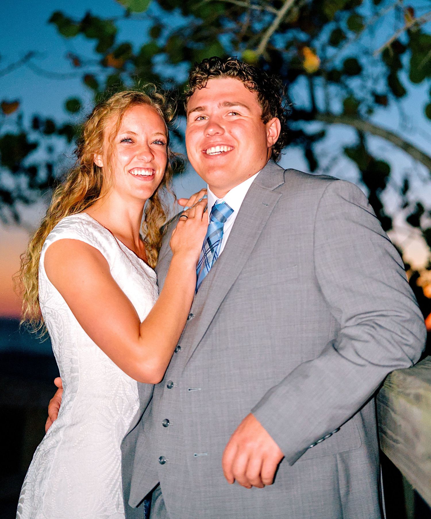 Happy couple posing outdoors at sunset, woman in white dress and man in gray suit with blue tie.
