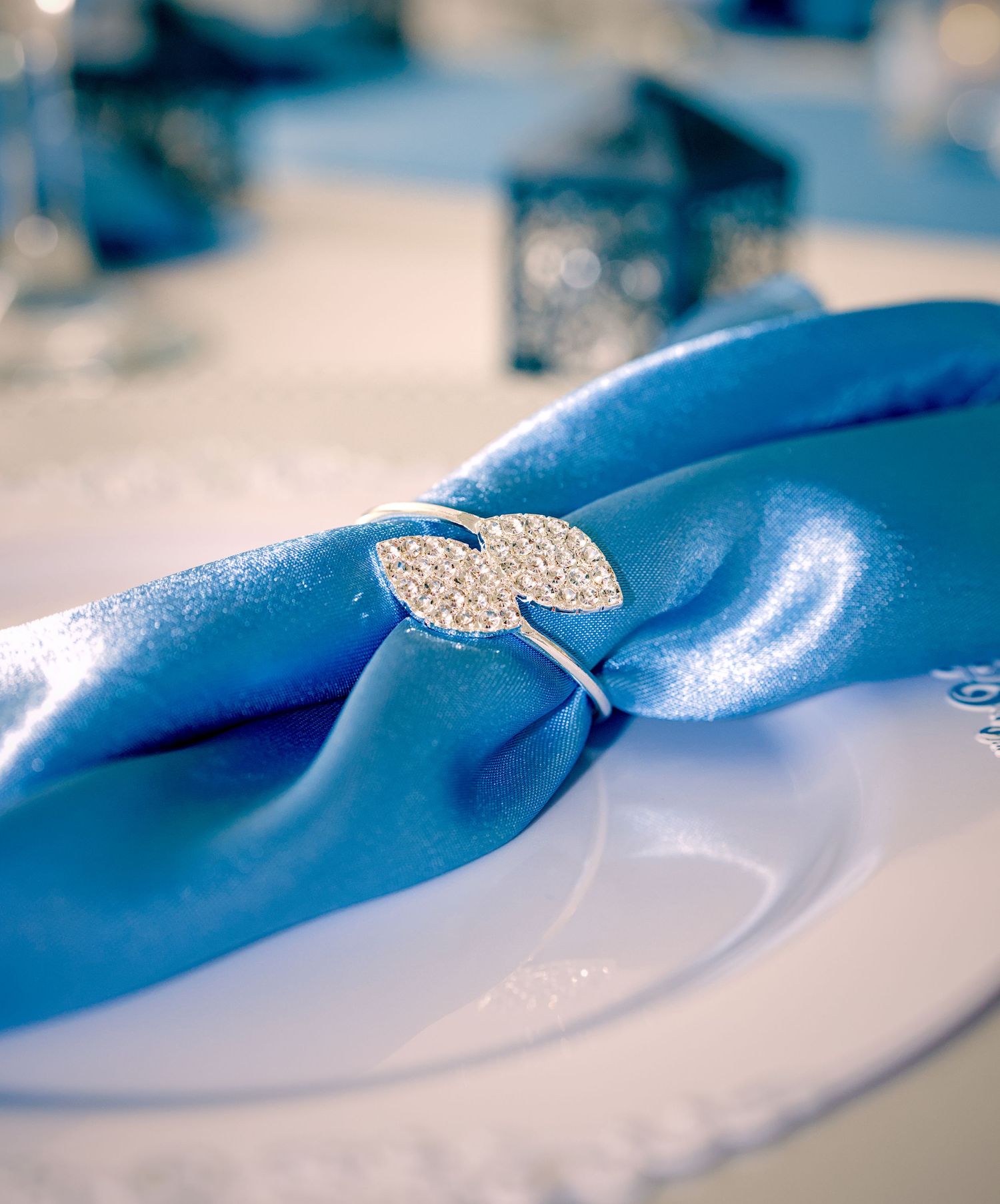 Elegant blue satin napkin with a sparkling rhinestone napkin ring on a white plate at a formal table setting.