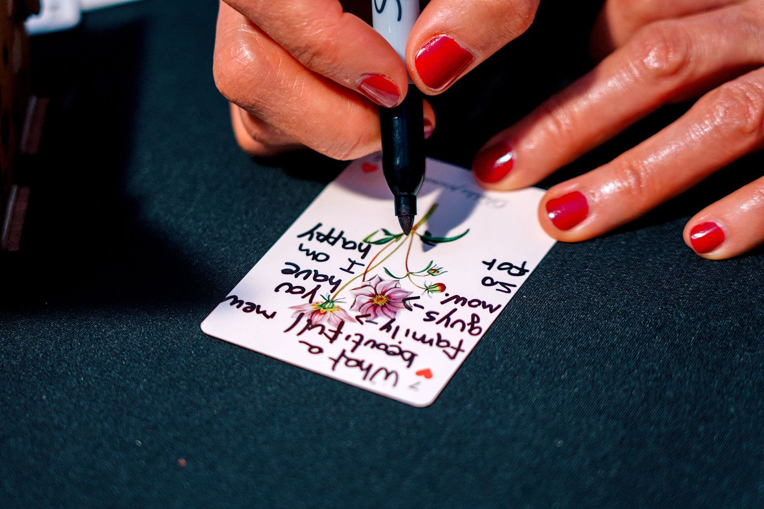 Close-up of a woman with red nails writing on a floral card with a black marker.