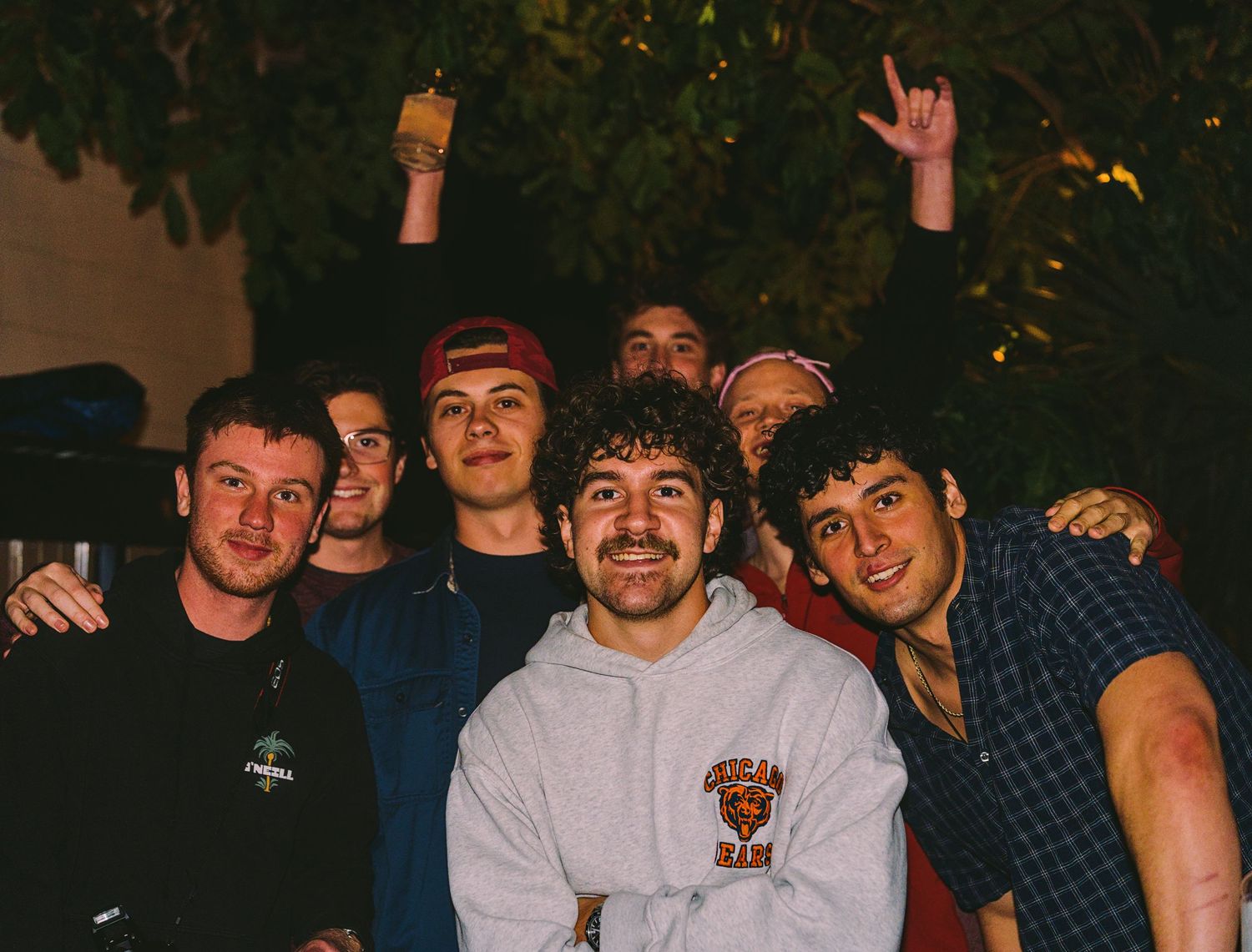 Group of young men smiling together outdoors at night, posing for a fun casual photo under trees.