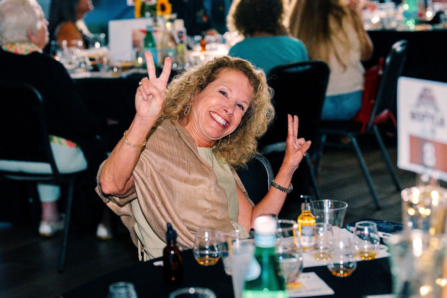 A smiling woman with curly hair making peace signs at an event table with drinks.