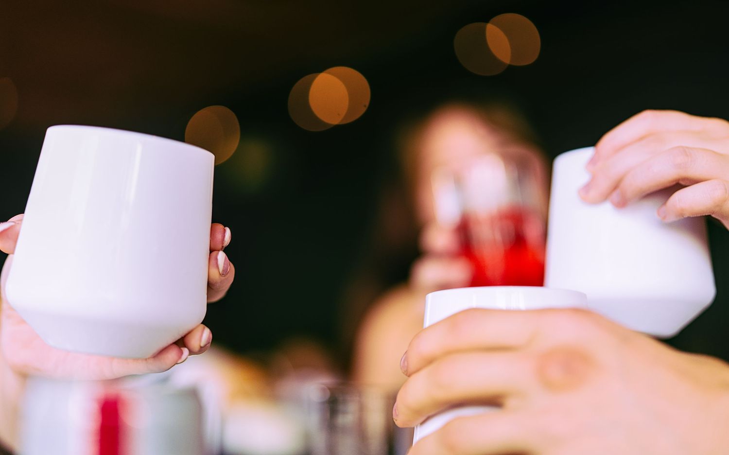 People clinking white ceramic mugs in a toast at a social gathering with bokeh lights in the background.