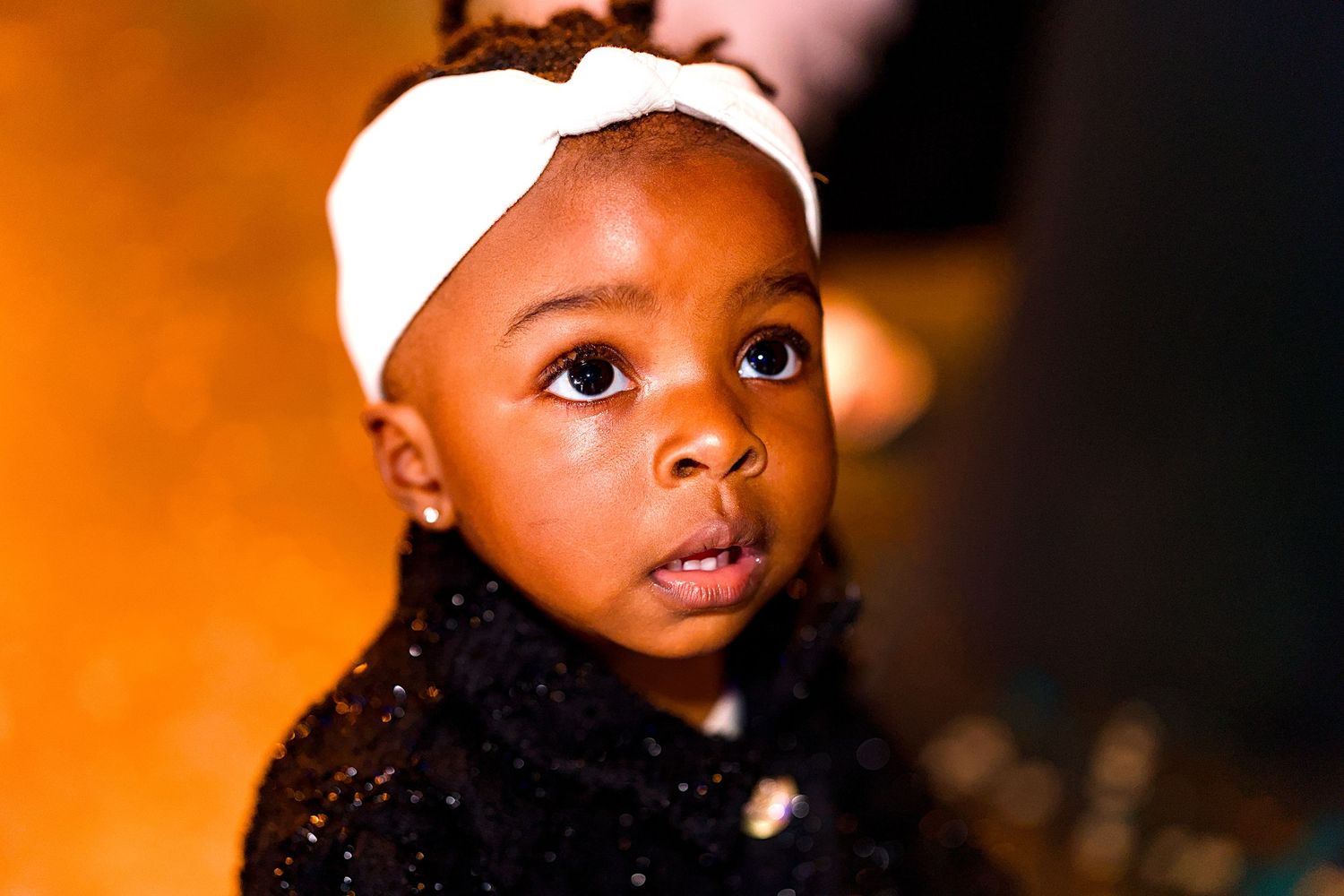 Adorable young Black girl wearing a white bow headband and black sparkly outfit looks up with big expressive eyes.