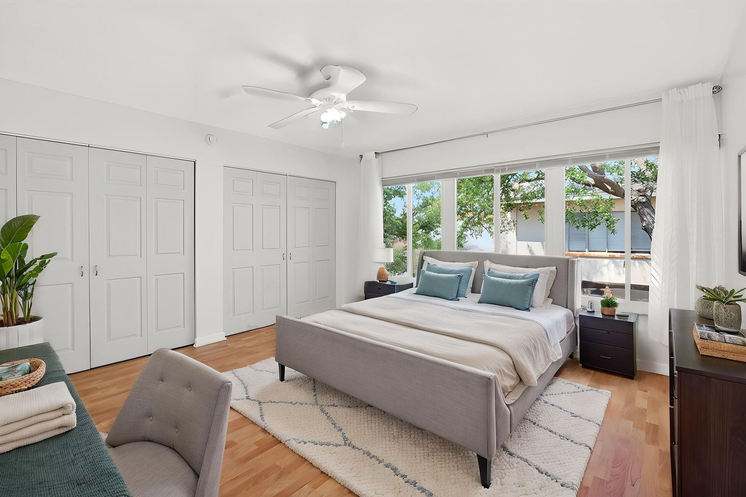 Modern bedroom with gray bed, hardwood floors, white walls, large windows, and indoor plants creating a bright airy space.