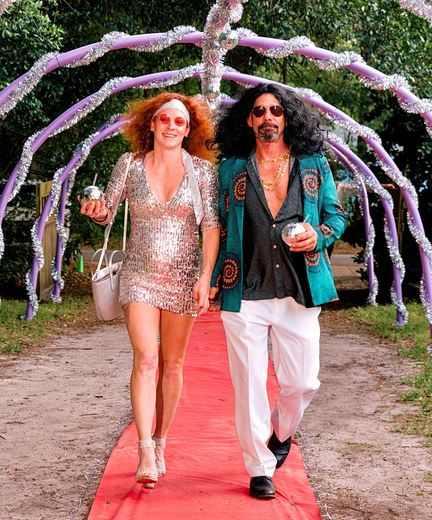 A couple in retro 70s disco outfits walk a red carpet under floral decorations at an outdoor event.