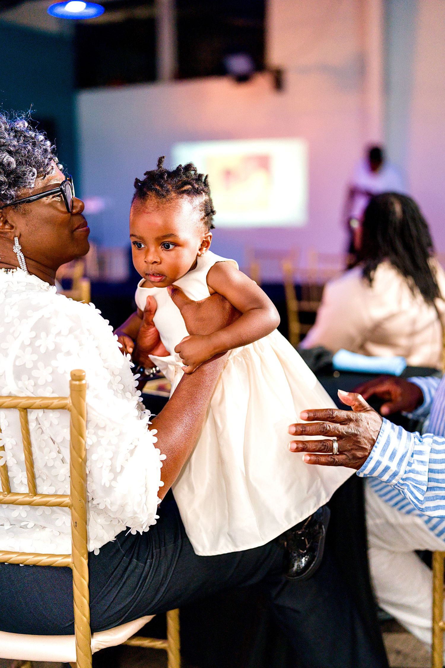 Elderly woman holds a baby girl in white dress at an elegant event with gold chairs and blue lighting.