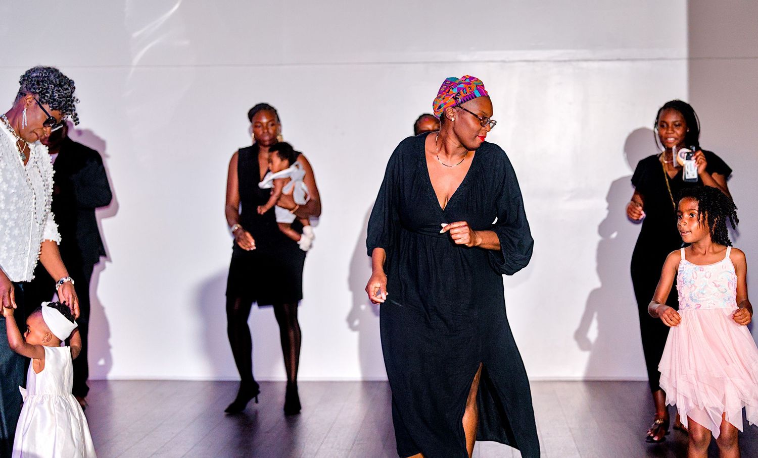 Group of Black adults and children dancing together at a social event in a bright white venue.