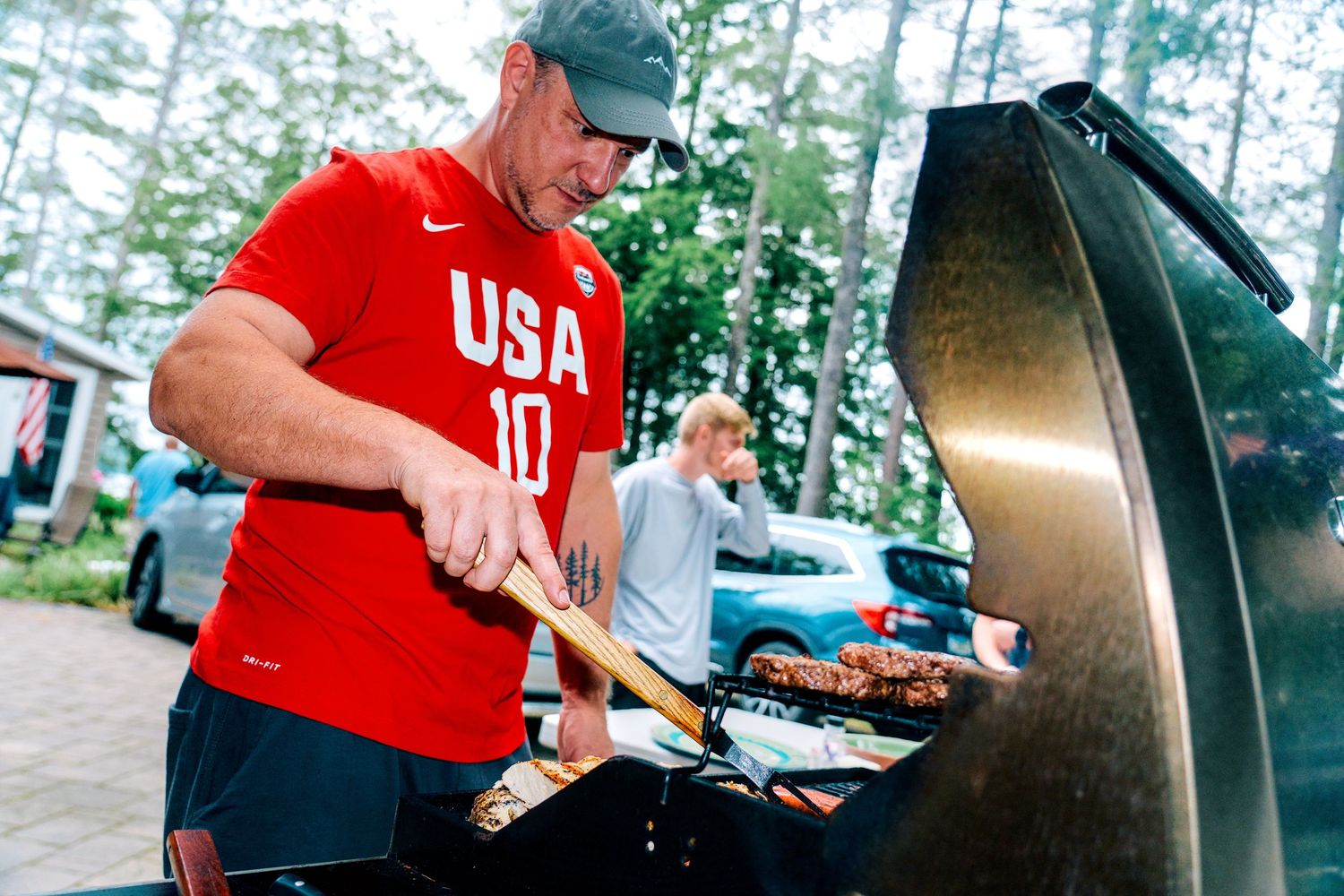Man in USA jersey grilling meat outdoors on a large black BBQ grill at a backyard gathering.