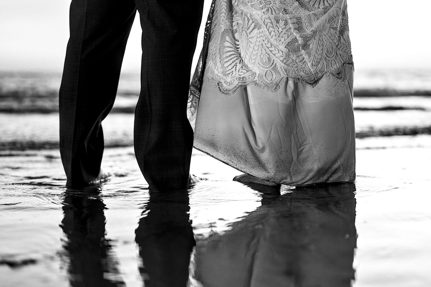 Black and white photo of bride and groom standing barefoot in ocean water, wedding dress trailing on wet sand.