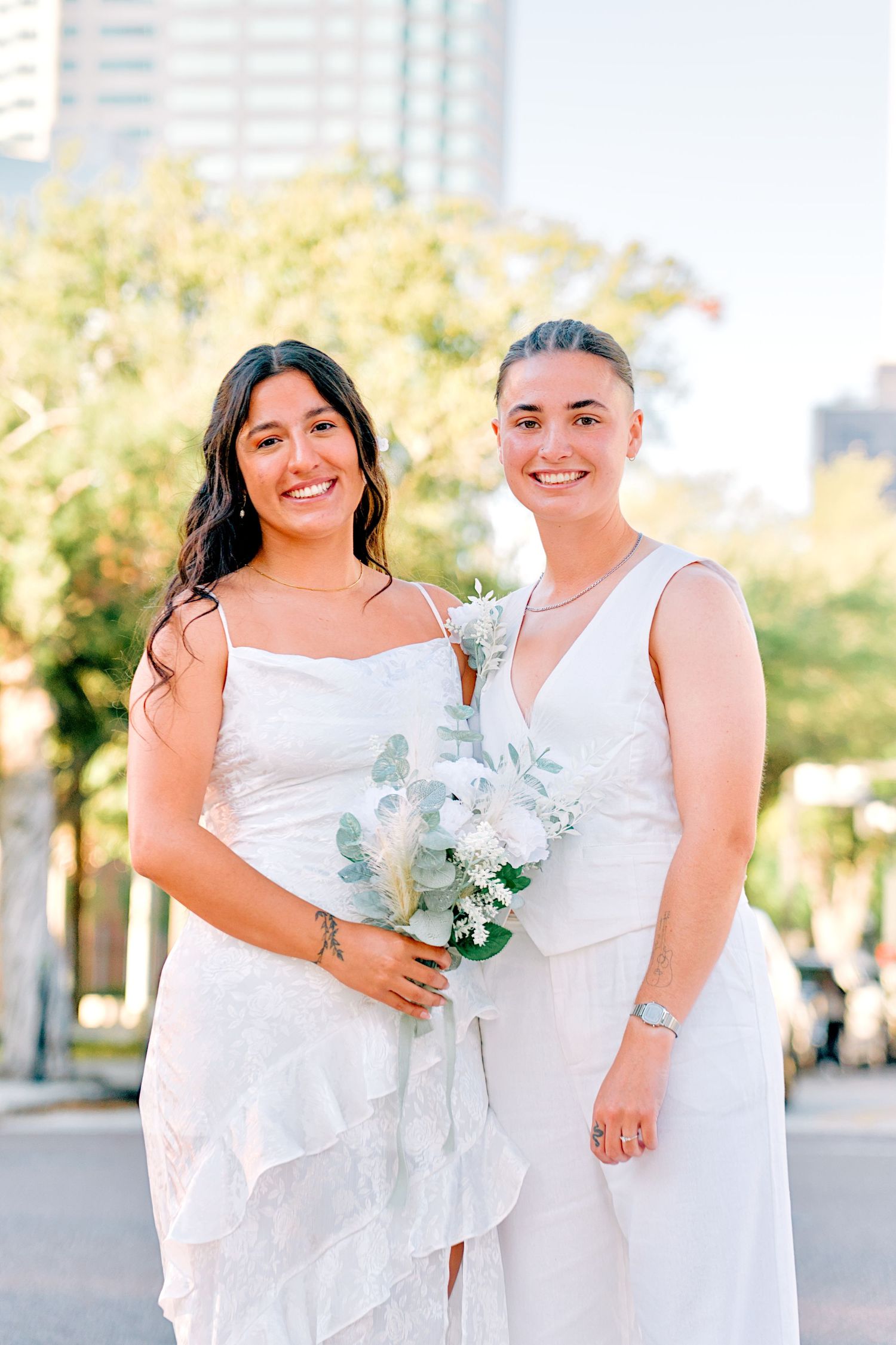 Two brides in white dresses smile together outdoors holding a white floral bouquet on their wedding day.