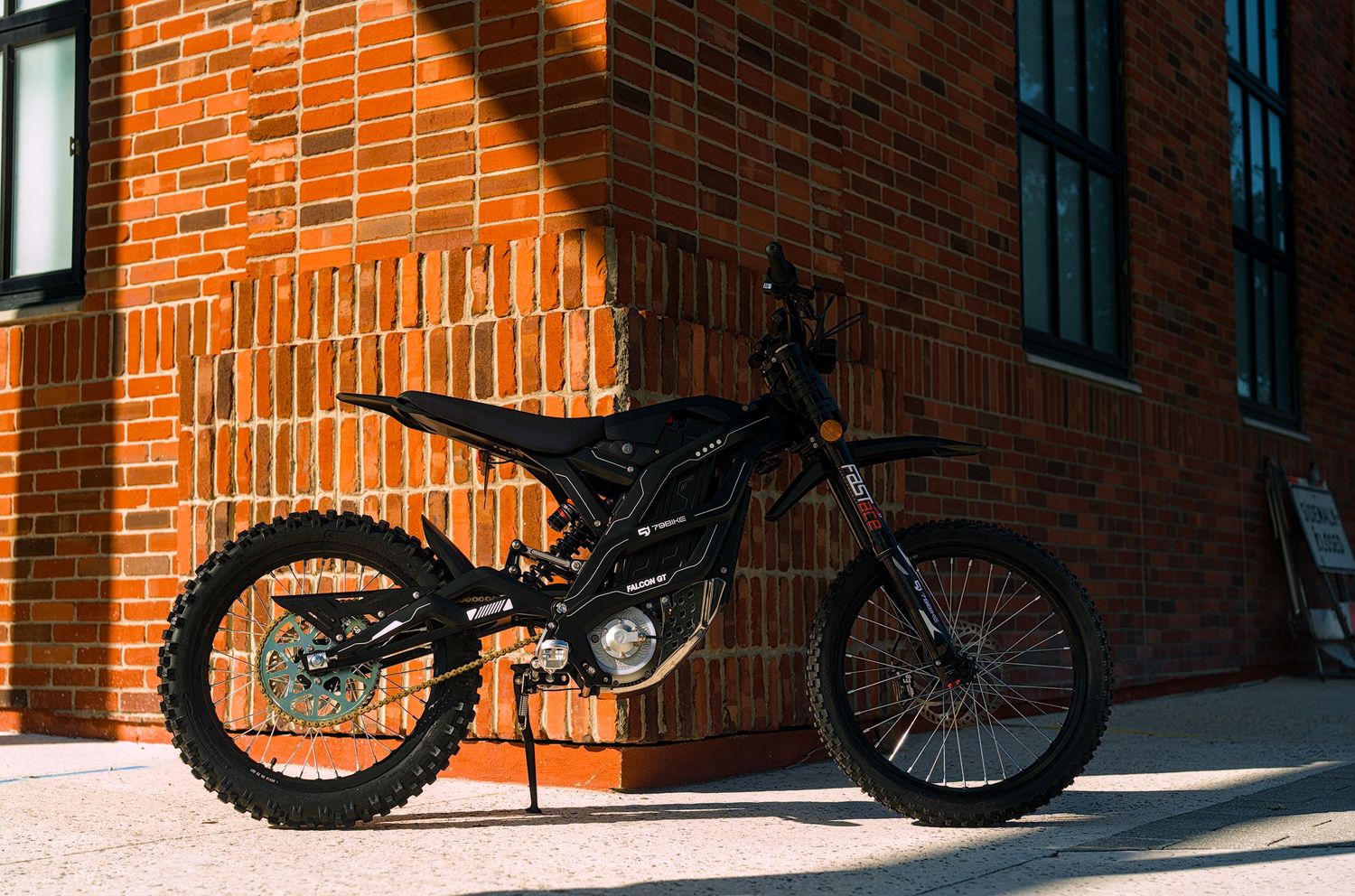 Black off-road dirt bike parked against a sunlit red brick wall, casting dramatic shadows on a sunny day.