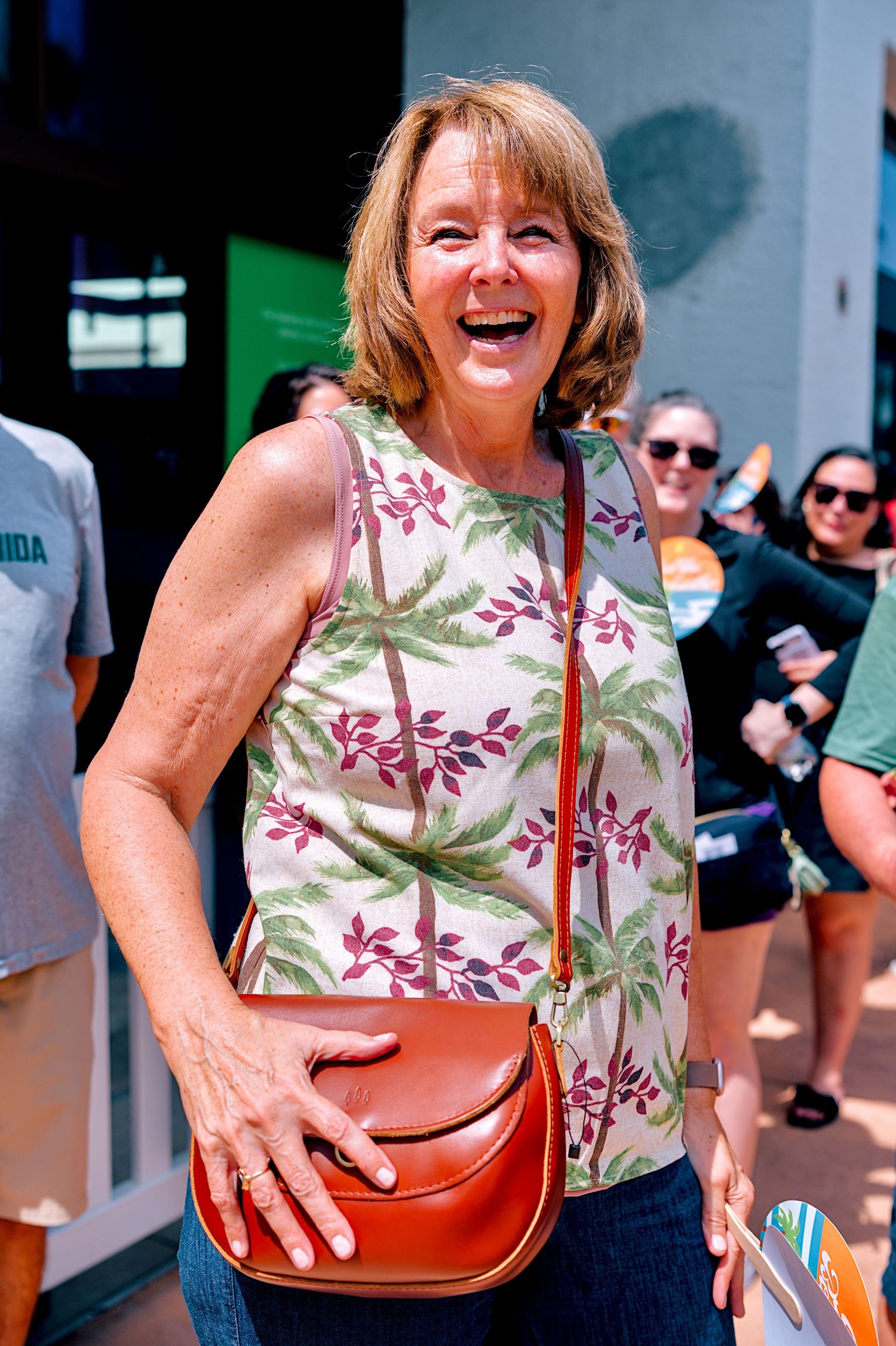 A smiling middle-aged woman in a floral palm tree top carries a brown crossbody bag outdoors in a sunny setting.