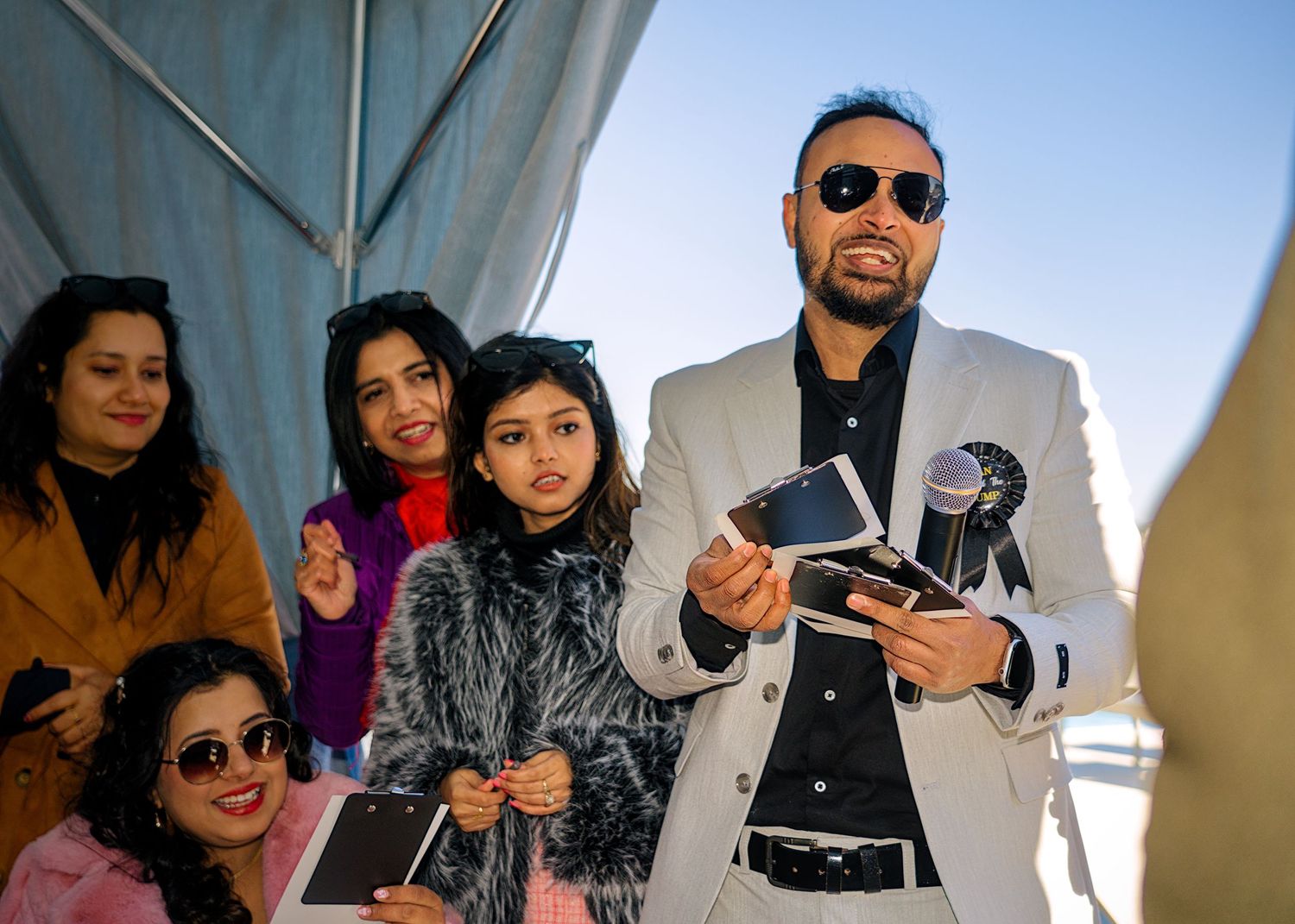A group of people wearing sunglasses and winter coats stand together outdoors on a sunny day.