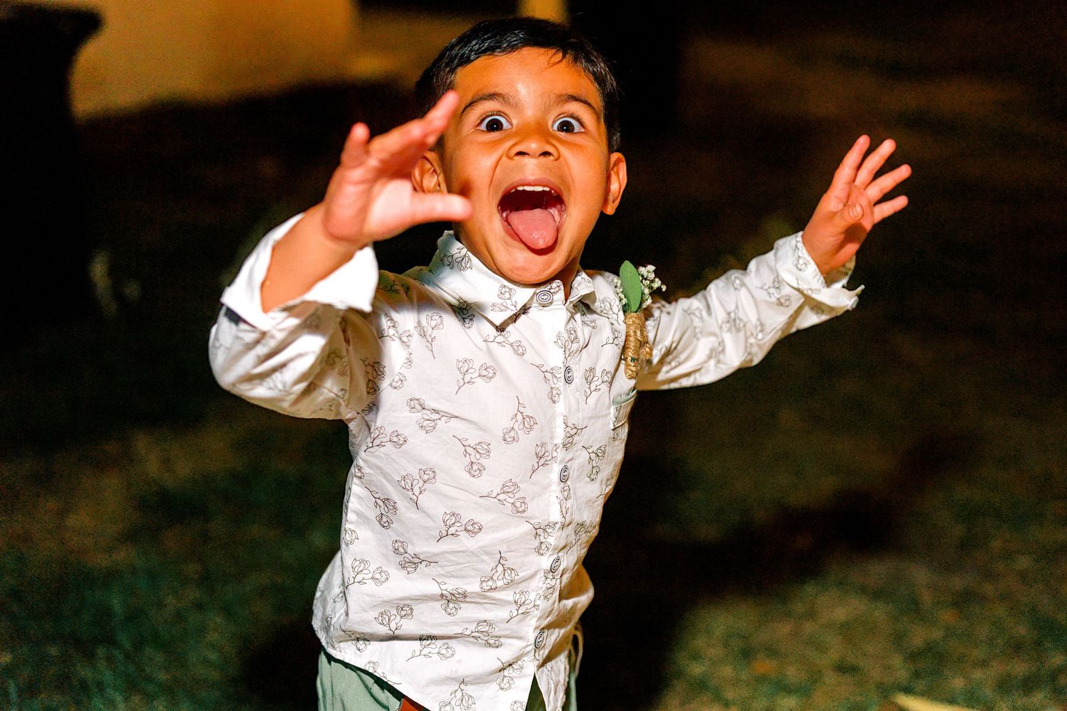Excited young boy in a white patterned shirt with a boutonniere, arms raised, mouth open wide at a wedding celebration.
