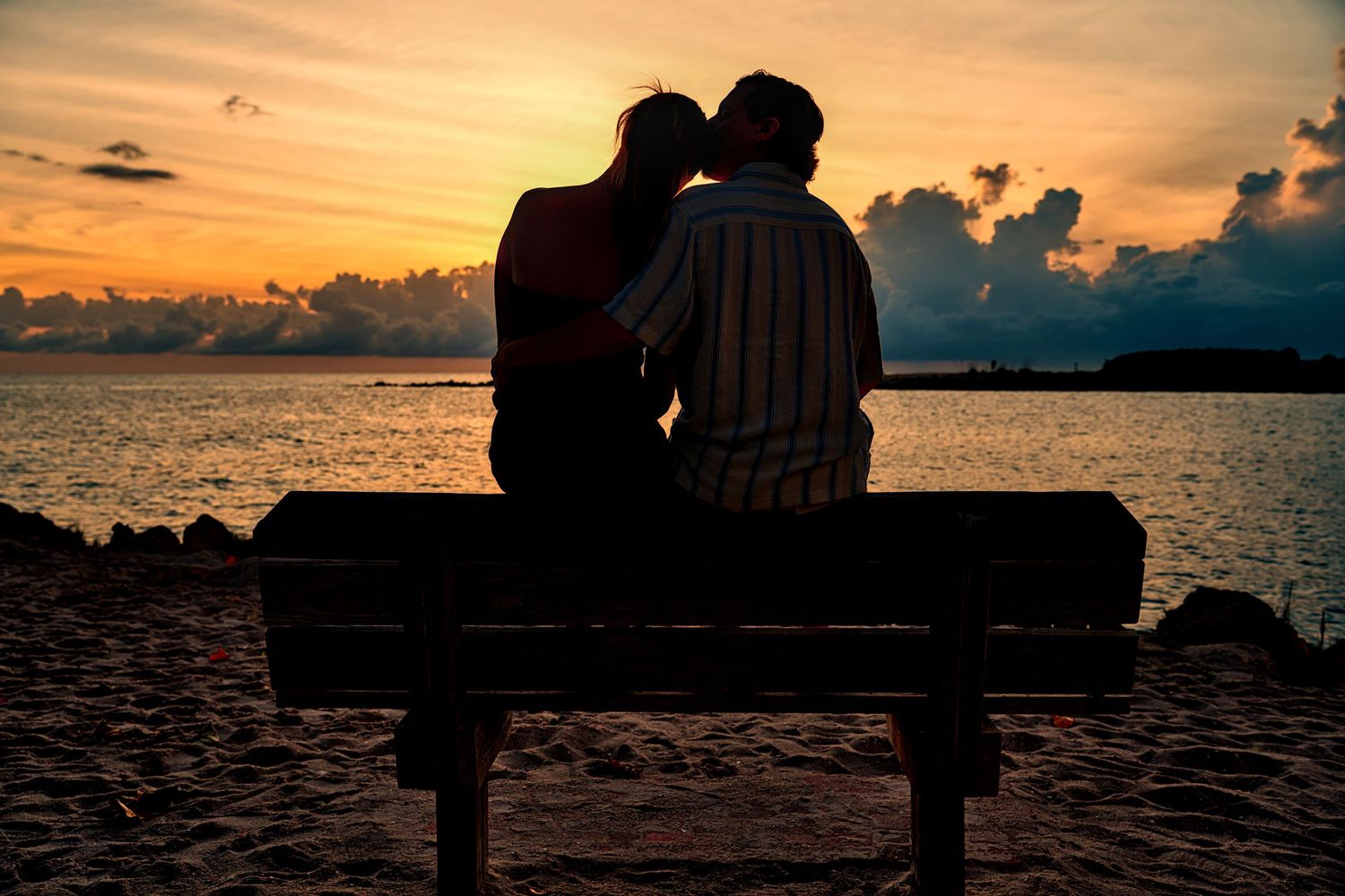 Silhouette of a romantic couple sitting on a bench sharing a kiss at a stunning ocean sunset.