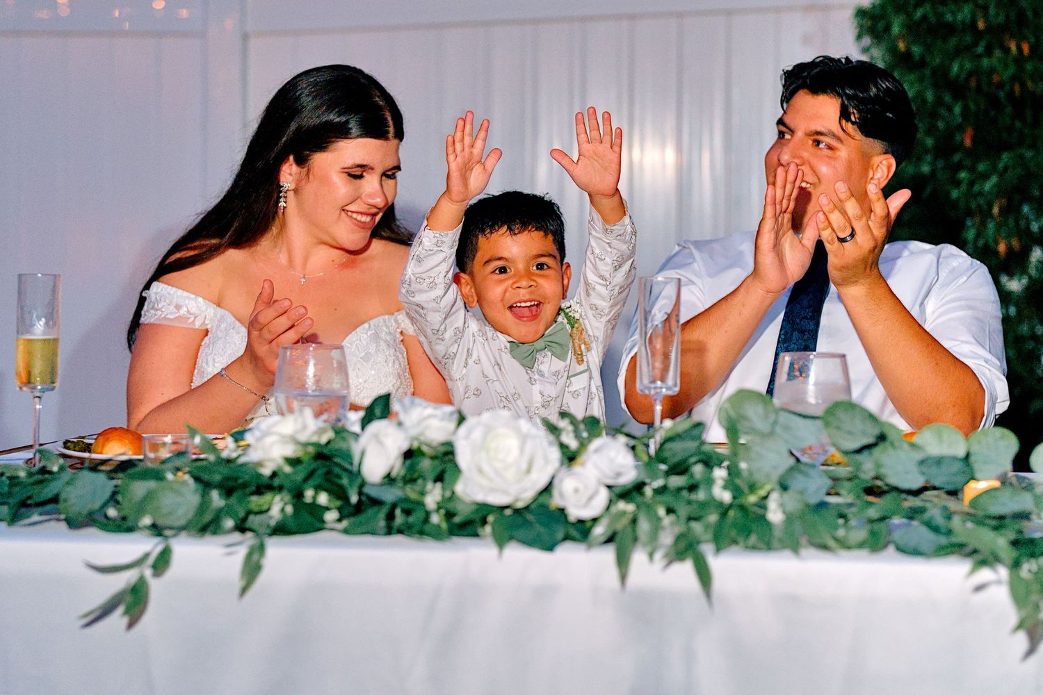 Bride, groom, and young boy celebrating joyfully at wedding reception head table with white florals.