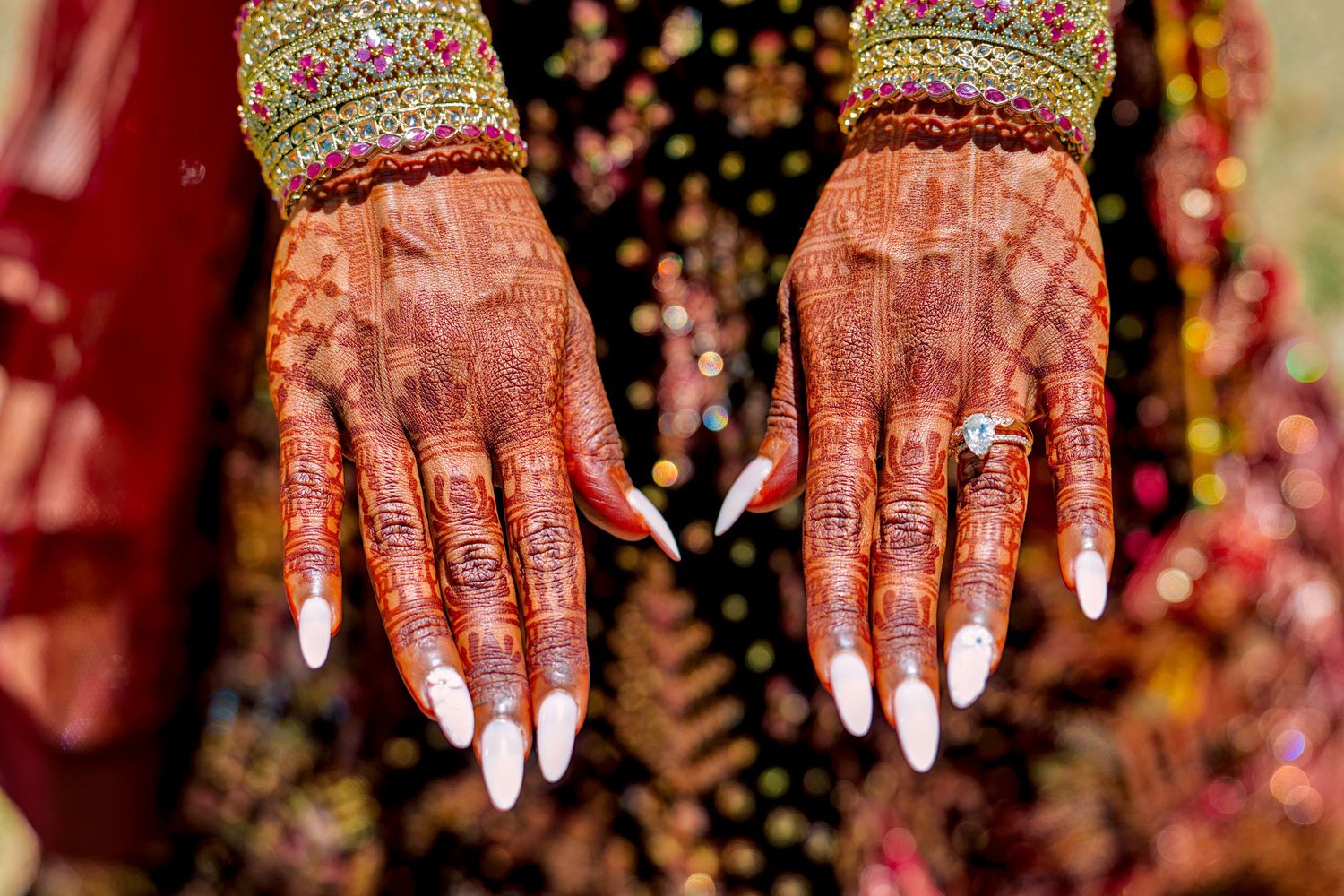 Close-up of bridal hands adorned with intricate henna mehndi designs and a diamond ring, with white nail extensions.