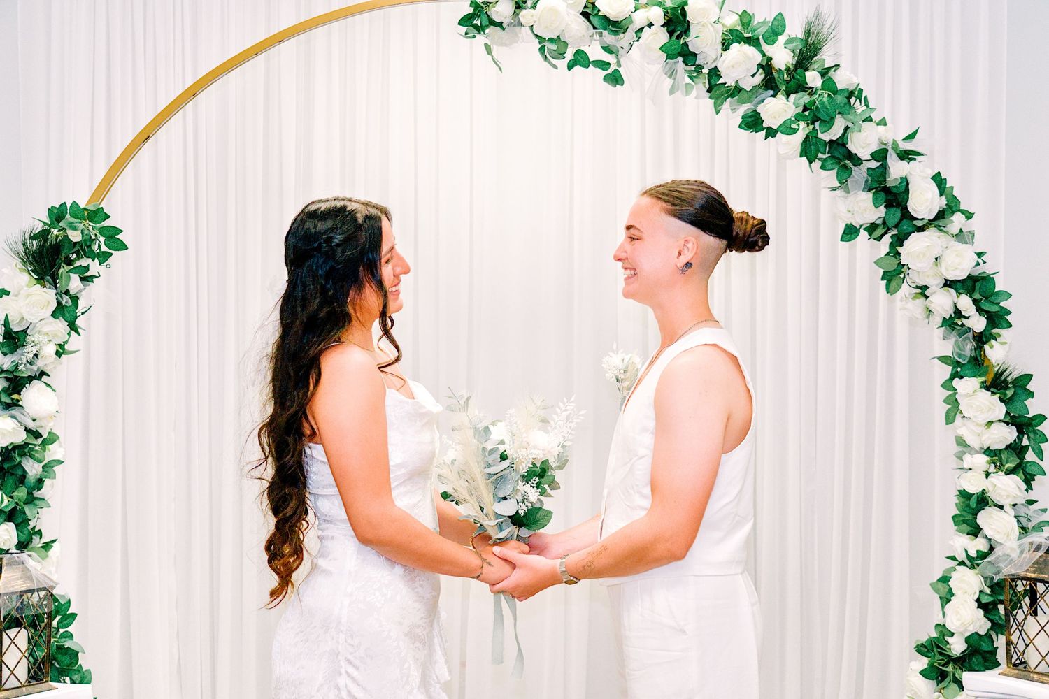 Two brides in white dresses hold hands smiling under a floral arch with white roses and greenery.
