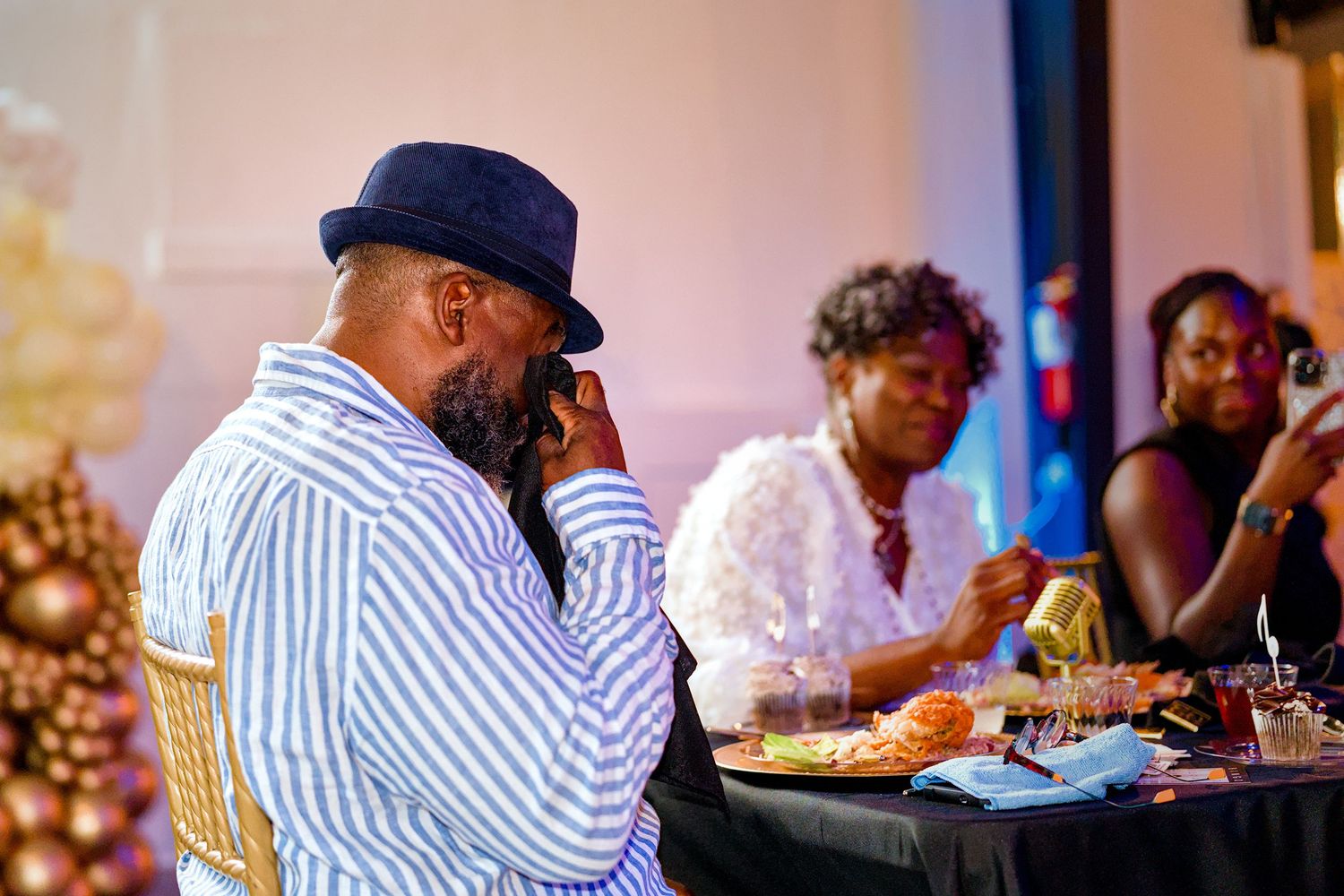 Man in blue striped shirt and hat sits at event table, thoughtfully touching his face while others dine nearby.