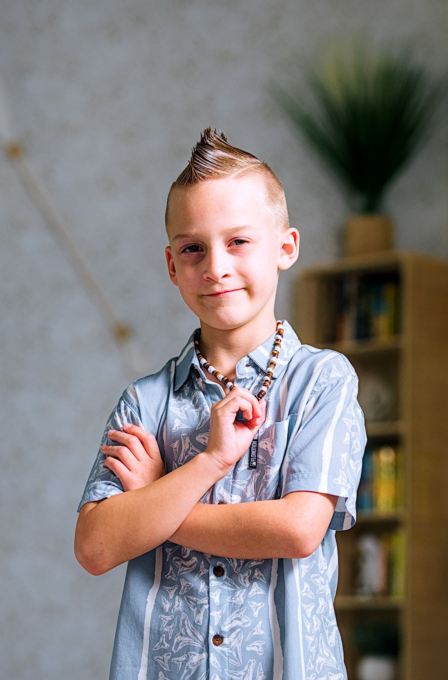 Young boy with a mohawk hairstyle wearing a blue patterned shirt and beaded necklace, posing with arms crossed.