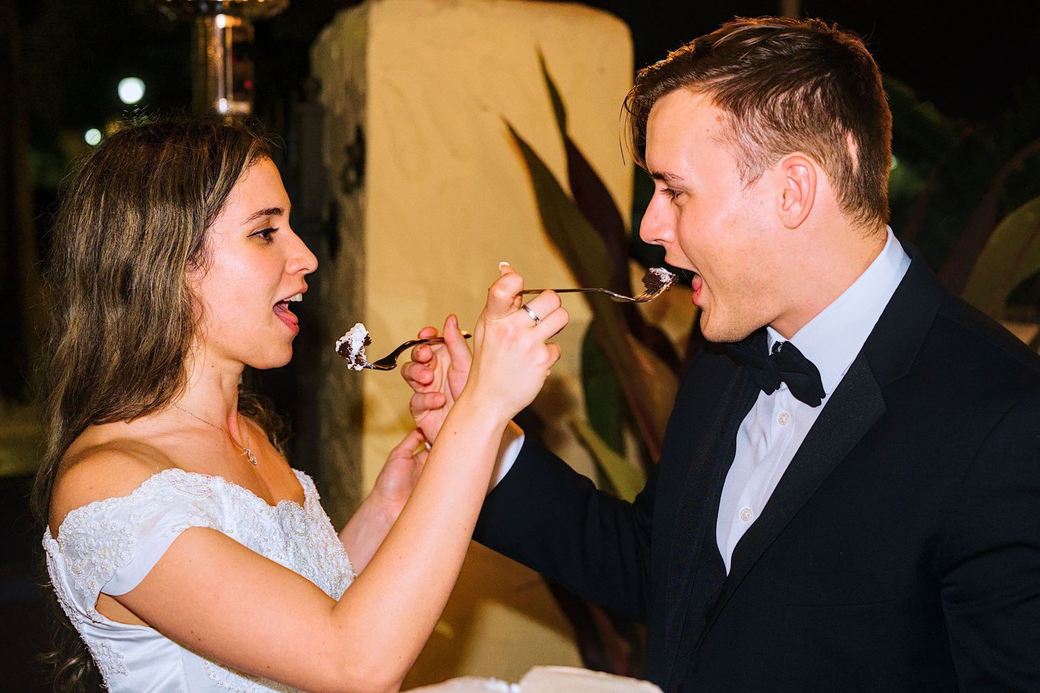 Bride and groom feeding each other wedding cake at their reception, smiling joyfully in formal attire.