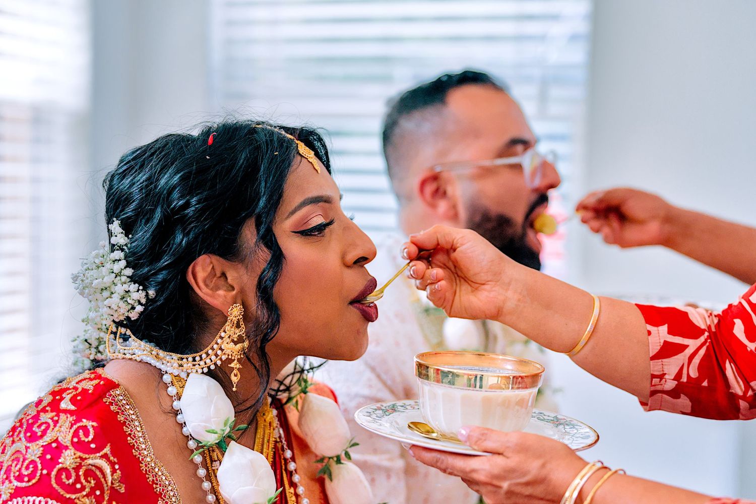Indian bride in red saree being fed during traditional wedding ceremony with groom in background.