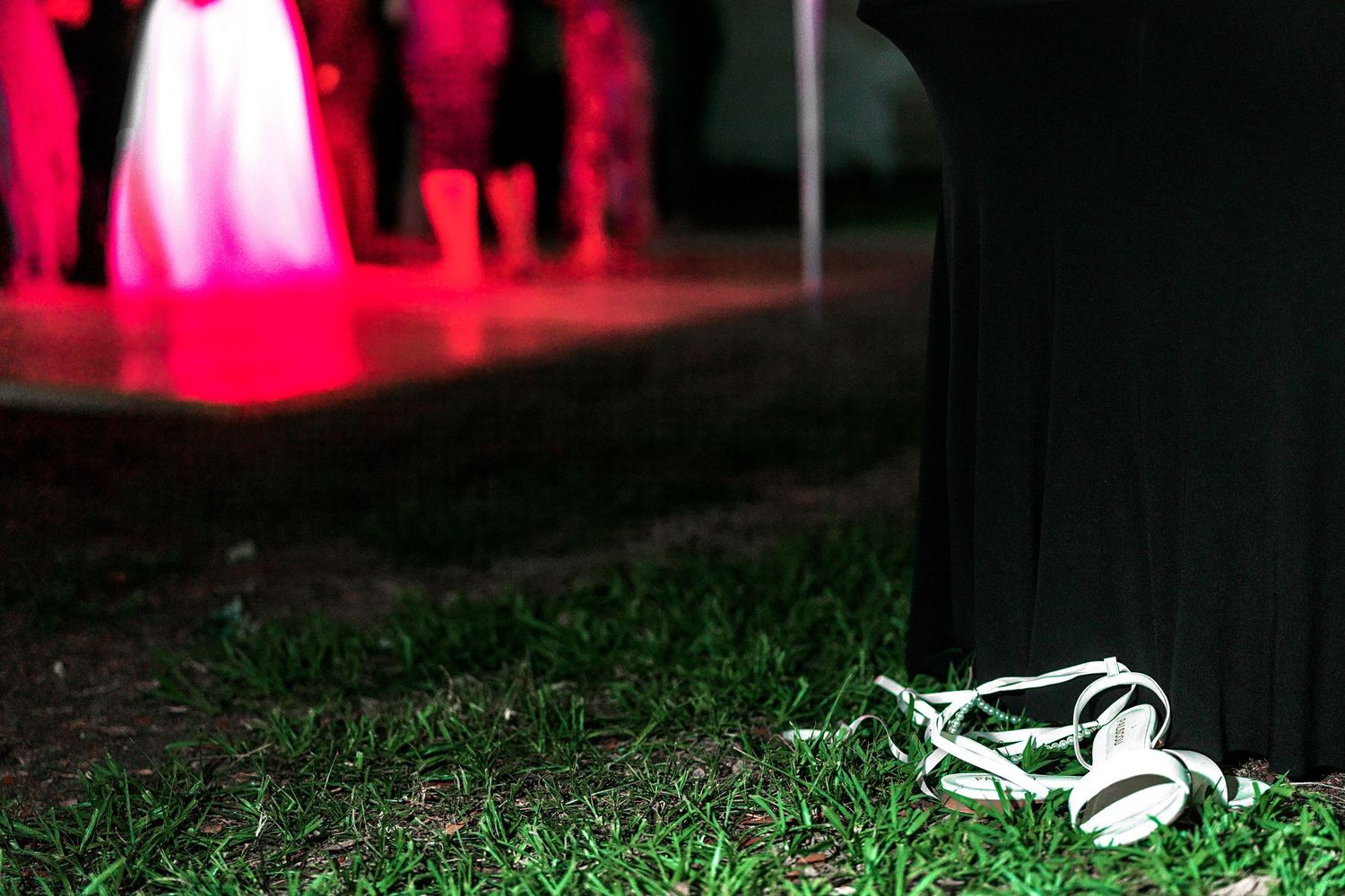White bridal heels left on green grass near dance floor with pink lights at outdoor wedding reception.